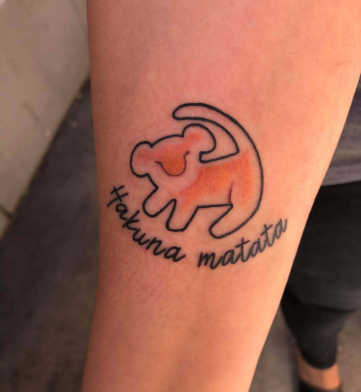 Tattoo of a stylized orange lion cub with "Hakuna Matata" text on an arm